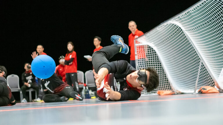 Male Japanese goalball athlete diving to keep a blue ball out of his net in the 2023 IBSA World Games goalball final.
